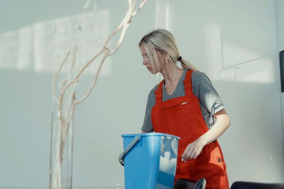 A woman in a red uniform cleans indoors with a blue bucket, ensuring a spotless environment.